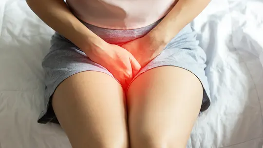 Die Person zeigt Anzeichen von Schmerzen, indem sie ihre Beine zusammenpresst und den Bauch mit beiden Händen hält.