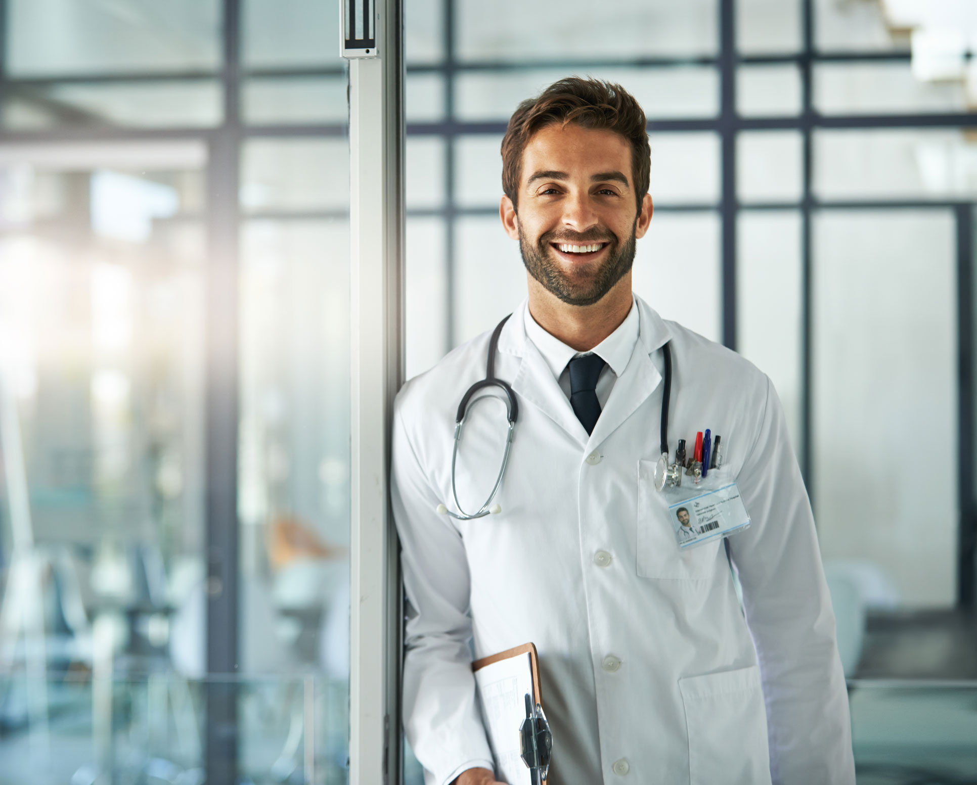 Arzt in weißen Kittel lächelt in die Kamera. Der Hintergrund ist unscharf und kühl gehalten.