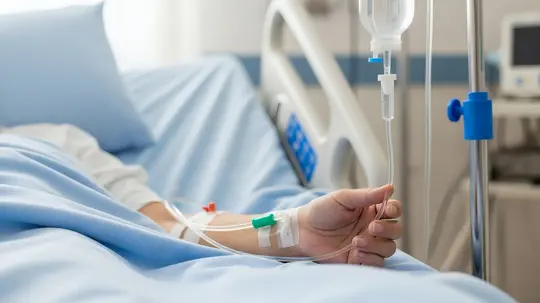 Eine Hand liegt auf einer Bettdecke, während ein Tropf an einem IV-Schlauch befestigt ist, was auf eine medizinische Behandlung hinweist.